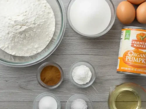 Top view of pumpkin cake ingredients on a wooden table including a bowl of flour, a bowl of sugar, eggs, canned pumpkin puree, vegetable oil, ground cinnamon, baking powder, baking soda, and salt