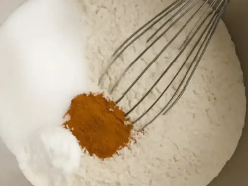 Stainless steel mixing bowl with flour, sugar, baking soda, and cinnamon being whisked together on a wooden table.