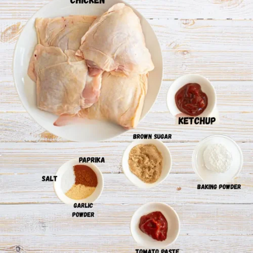 Raw Chicken Thighs with Seasoning Ingredients for Marinade Preparation Overhead shot of raw bone-in, skin-on chicken thighs on a white plate surrounded by small bowls of ingredients including ketchup, tomato paste, brown sugar, baking powder, paprika, garlic powder, and salt on a light wooden background.
